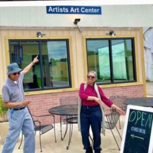 Two smiling individuals enthusiastically welcome visitors outside the Artists Art Center, a creative space for artists and art lovers. A chalkboard sign reads "OPEN – Come on in," inviting guests to explore the center.
