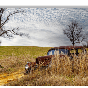 Photograph of an abandoned Hudson car rusting in a grassy field under a cloudy sky.