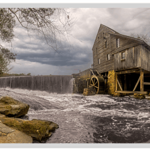 Fine art photo of Yates Mill with rushing water over the stone dam under stormy skies.