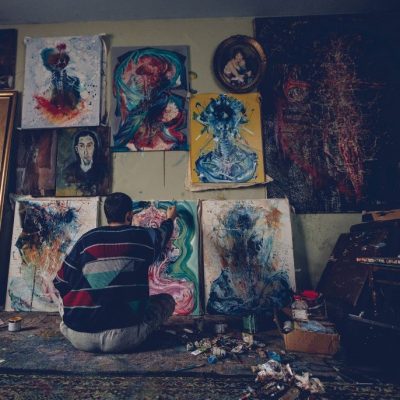 An artist sits on the floor of a dimly lit studio, painting an abstract canvas while surrounded by expressive, colorful artworks on the walls and floor.