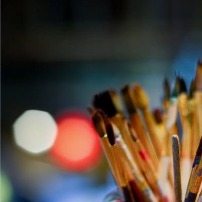 A smiling female artist wearing a yellow apron sits on a stool in a studio, surrounded by vibrant abstract paintings, paint cans, and brushes